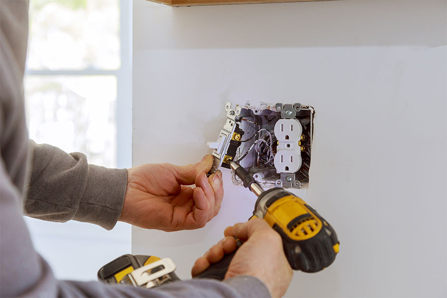 Person installs or repairs a light switch next to a power outlet using a cordless drill; internal wires are visible.