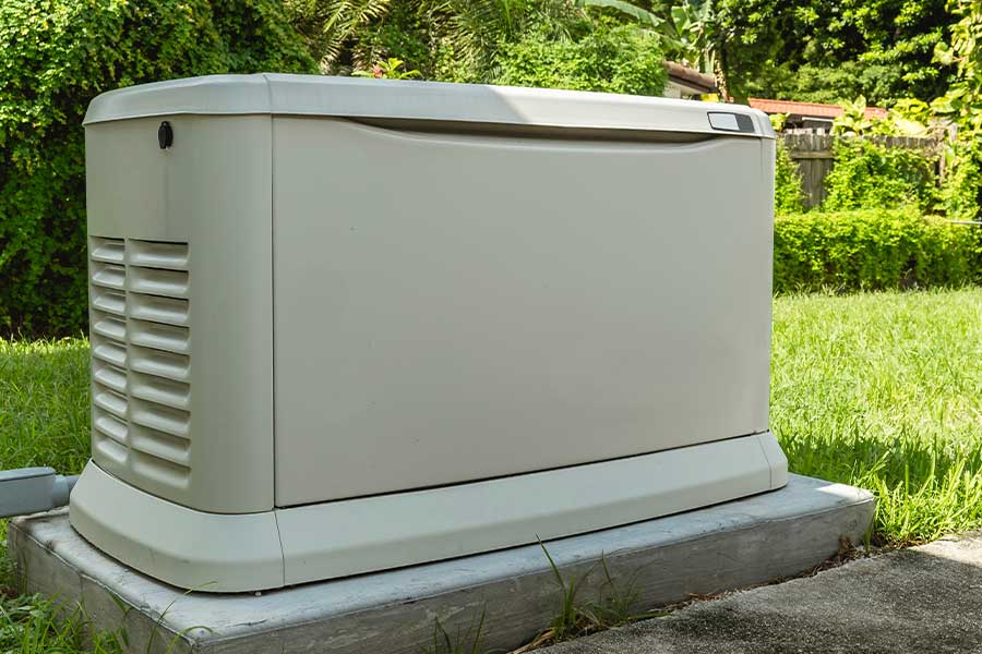 A large outdoor standby generator sits on a concrete platform in a grassy yard, surrounded by trees and bushes.