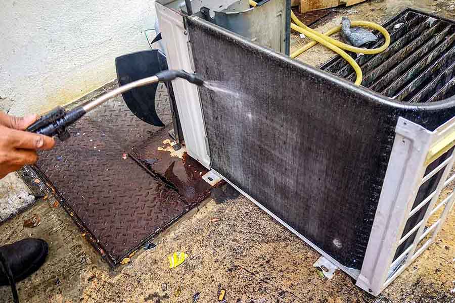 A person uses a pressure washer to clean the coils of an outdoor air conditioning unit, with water and debris on the ground.