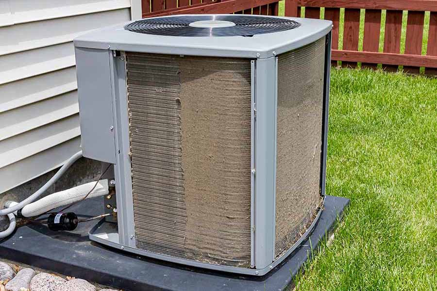Outdoor air conditioning unit with dirty condenser coils placed on a concrete pad next to a house, surrounded by grass and a wooden fence.