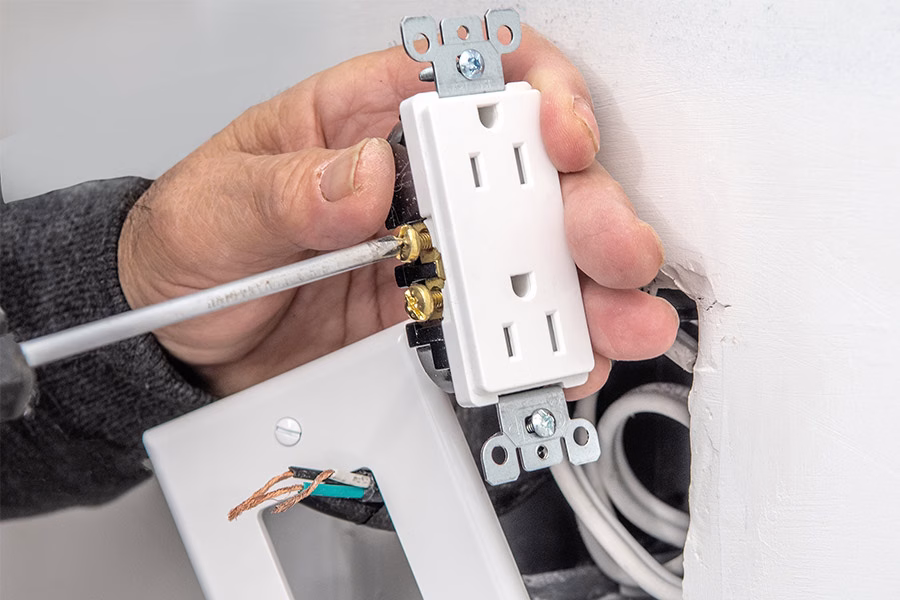 A person installs a standard electrical outlet in a wall, tightening a screw with a screwdriver. The cover plate and wires are visible.