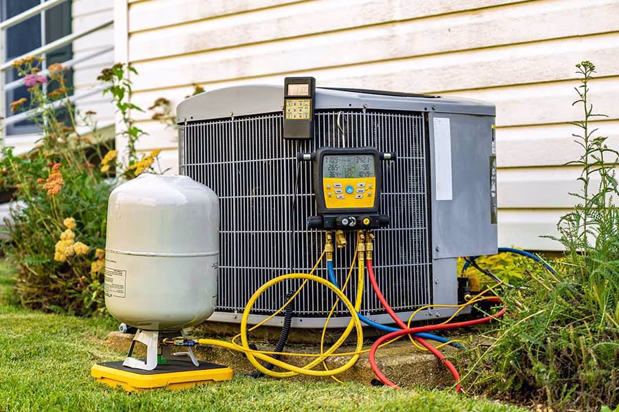 An air conditioning unit outside a house with diagnostic gauges and hoses attached, and a refrigerant tank on the ground nearby.